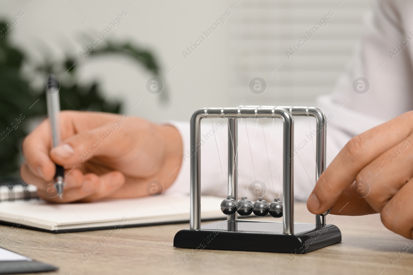 Photo of Man playing with Newton's cradle and writing something in notebook at wooden table, closeup. Physics law of energy conservation