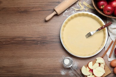 Photo of Flat lay composition with raw dough, fork and ingredients on wooden table, space for text. Baking apple pie