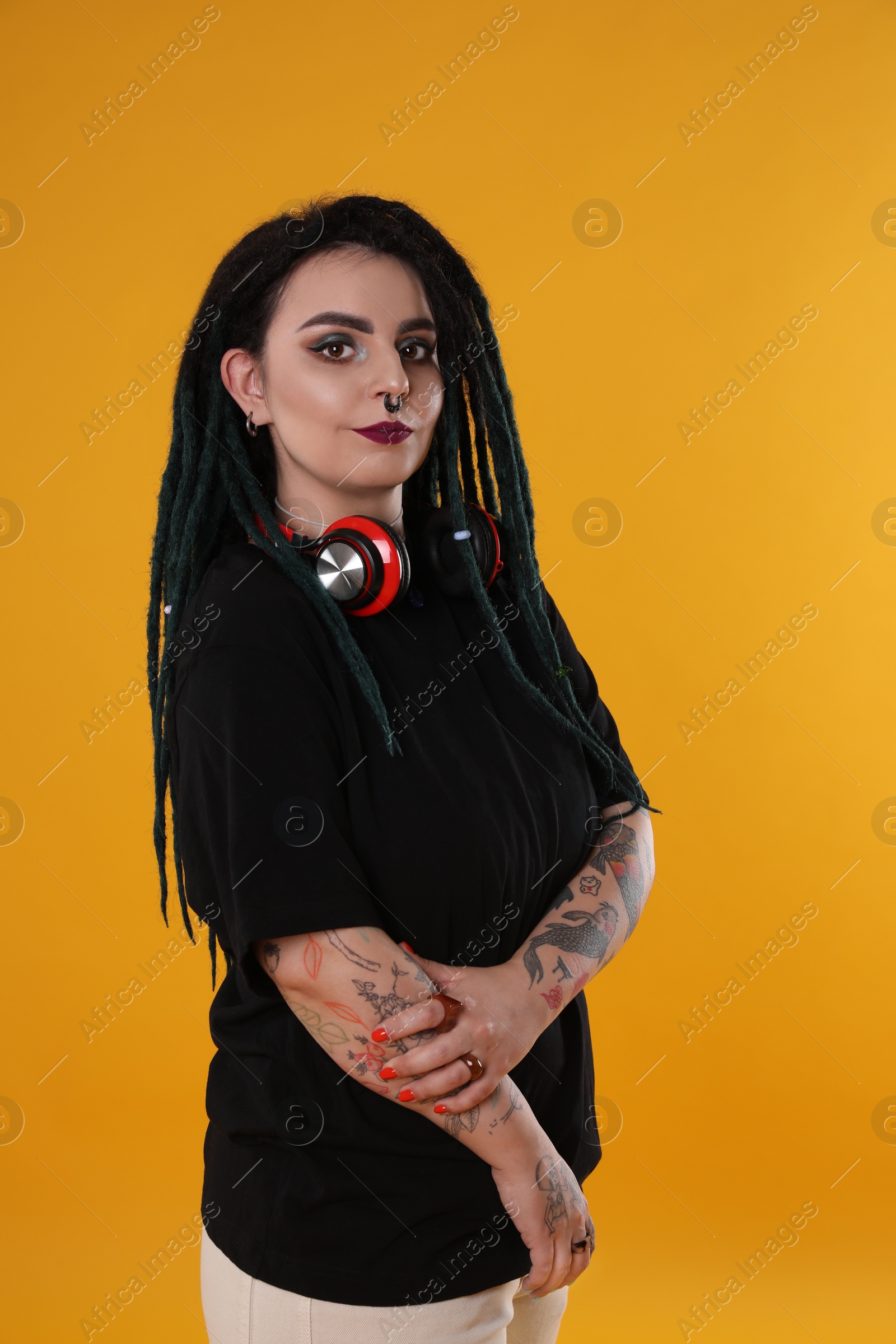 Photo of Beautiful young woman with tattoos on arms, nose piercing and dreadlocks against yellow background