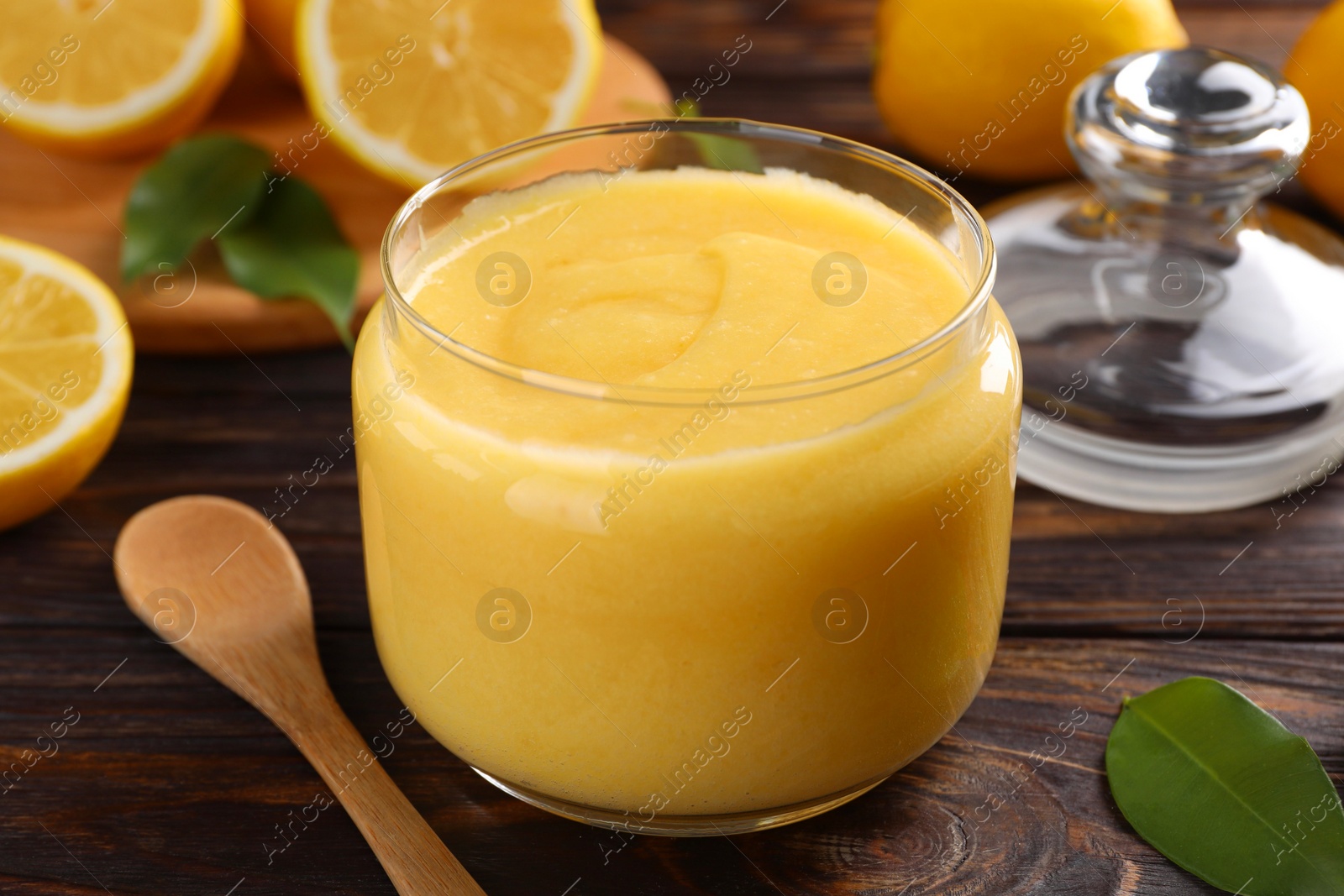 Photo of Delicious lemon curd in glass jar, fresh citrus fruits and spoon on wooden table, closeup