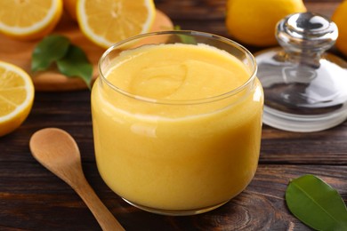 Delicious lemon curd in glass jar, fresh citrus fruits and spoon on wooden table, closeup