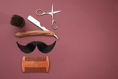 Photo of Artificial moustache and barber tools on burgundy background, flat lay. Space for text