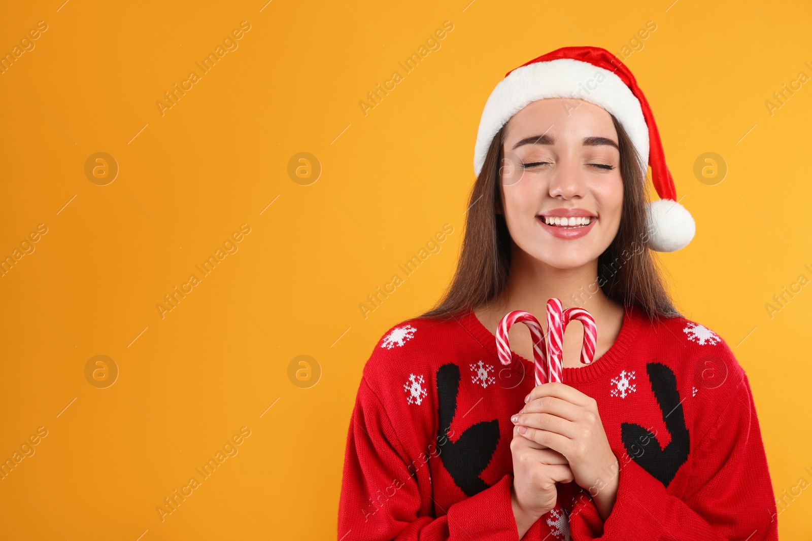 Photo of Young woman in Christmas sweater and Santa hat holding candy canes on yellow background, space for text