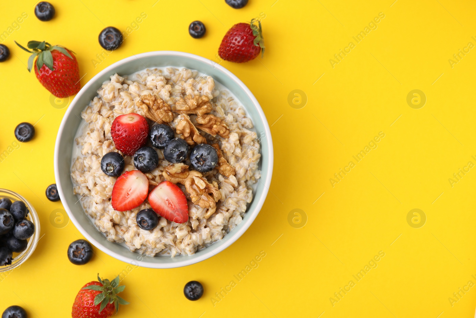 Photo of Tasty oatmeal with strawberries, blueberries and walnuts in bowl surrounded by ingredients on yellow background, flat lay. Space for text