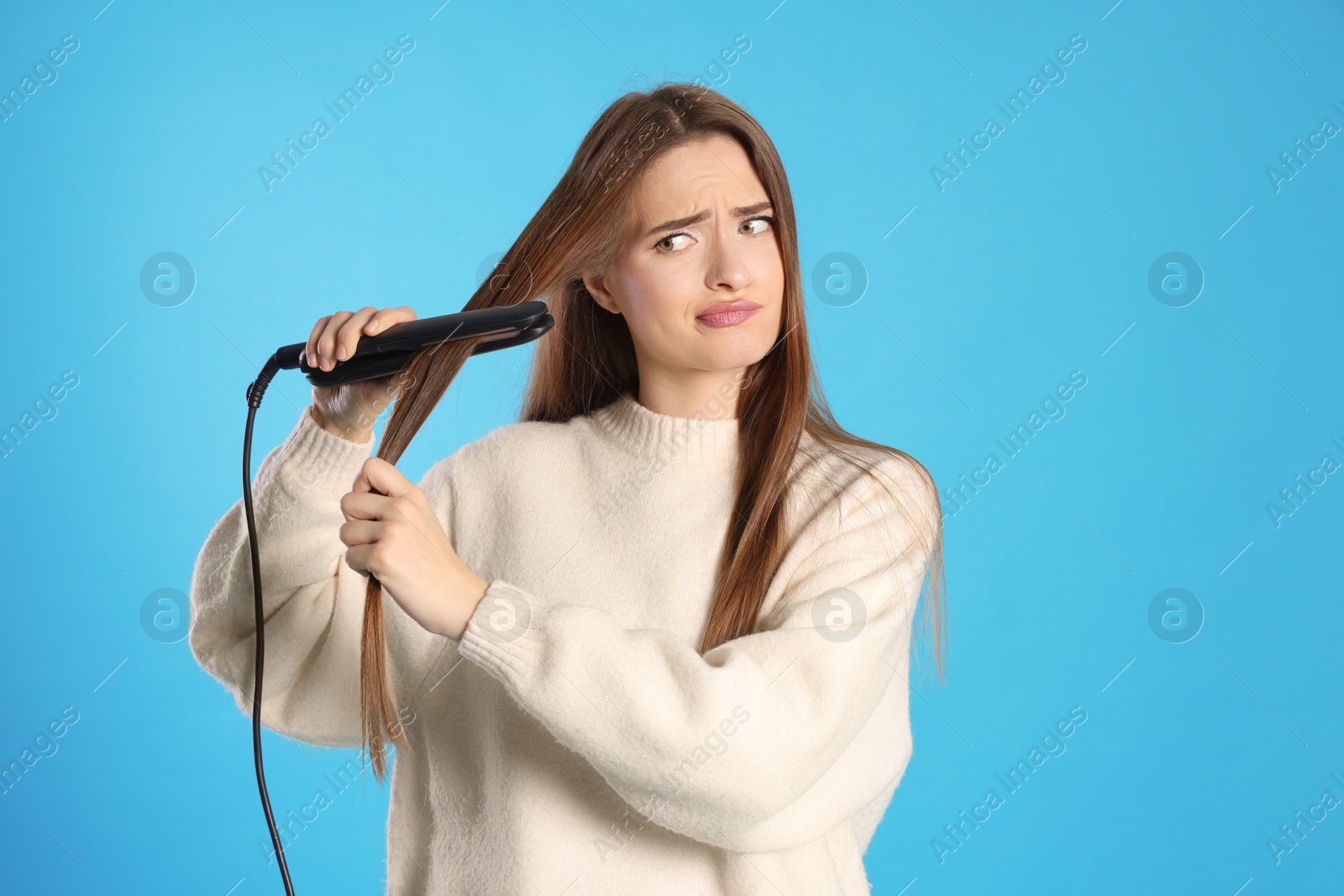 Photo of Upset young woman with flattening iron on light blue background. Hair damage