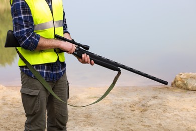 Man with hunting rifle wearing safety vest near lake outdoors, closeup