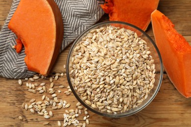 Dry pearl barley in bowl and pieces of pumpkin on wooden table, flat lay