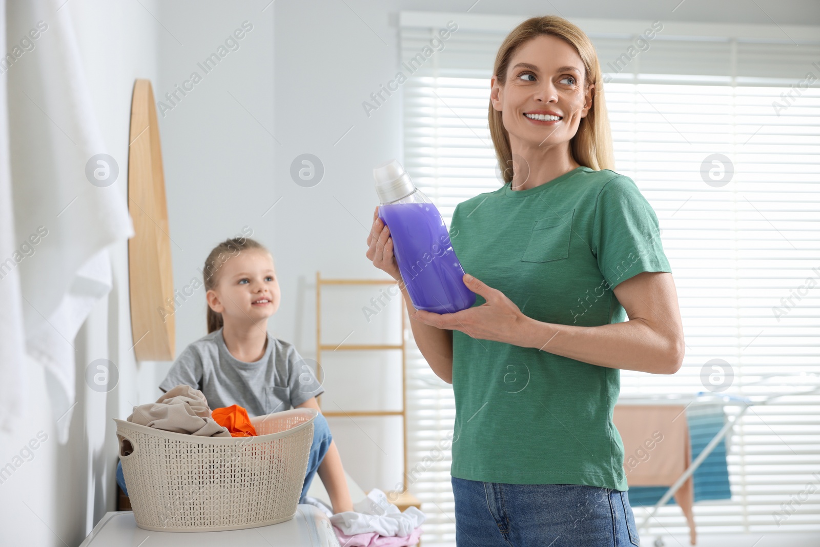 Photo of Mother holding fabric softener and daughter sitting near basket with dirty clothes in bathroom