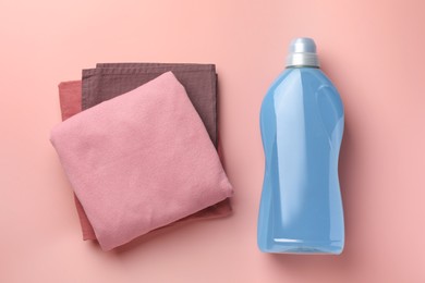 Photo of Bottle of fabric softener and stacked clean clothes on pink background, flat lay