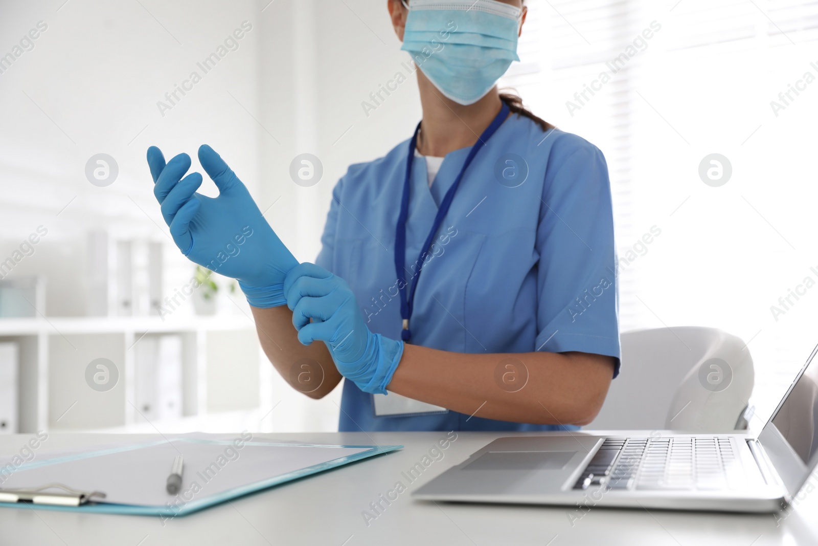 Photo of Doctor in protective mask putting on medical gloves at table in office, closeup