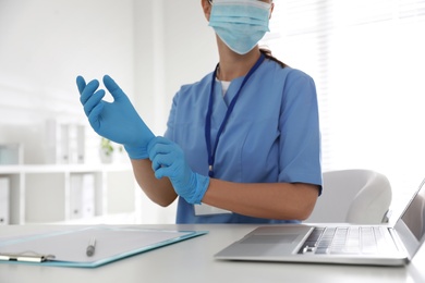 Doctor in protective mask putting on medical gloves at table in office, closeup