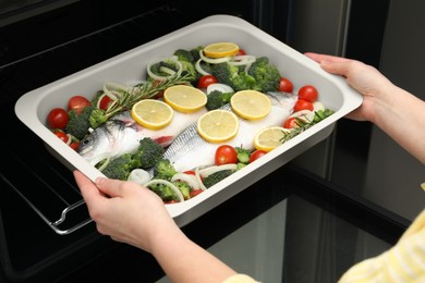 Photo of Woman putting baking dish with raw fish and vegetables into oven, closeup