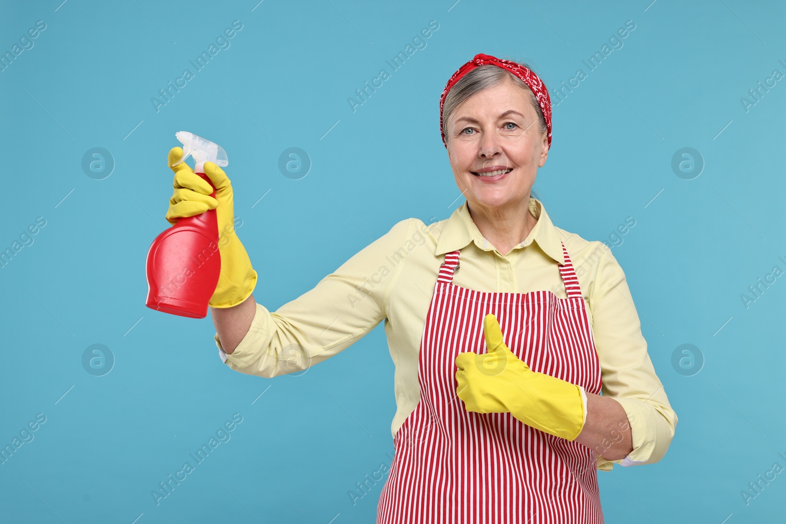 Photo of Happy housewife with spray bottle showing thumbs up on light blue background