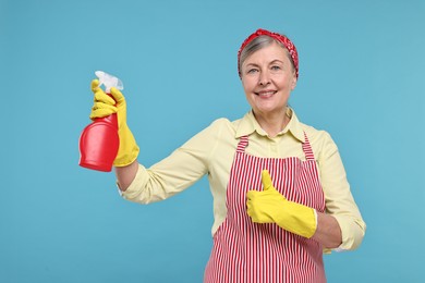 Happy housewife with spray bottle showing thumbs up on light blue background