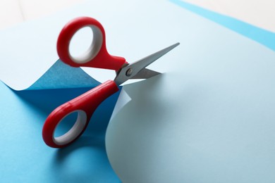 Photo of Red scissors cutting light blue paper on white background, closeup