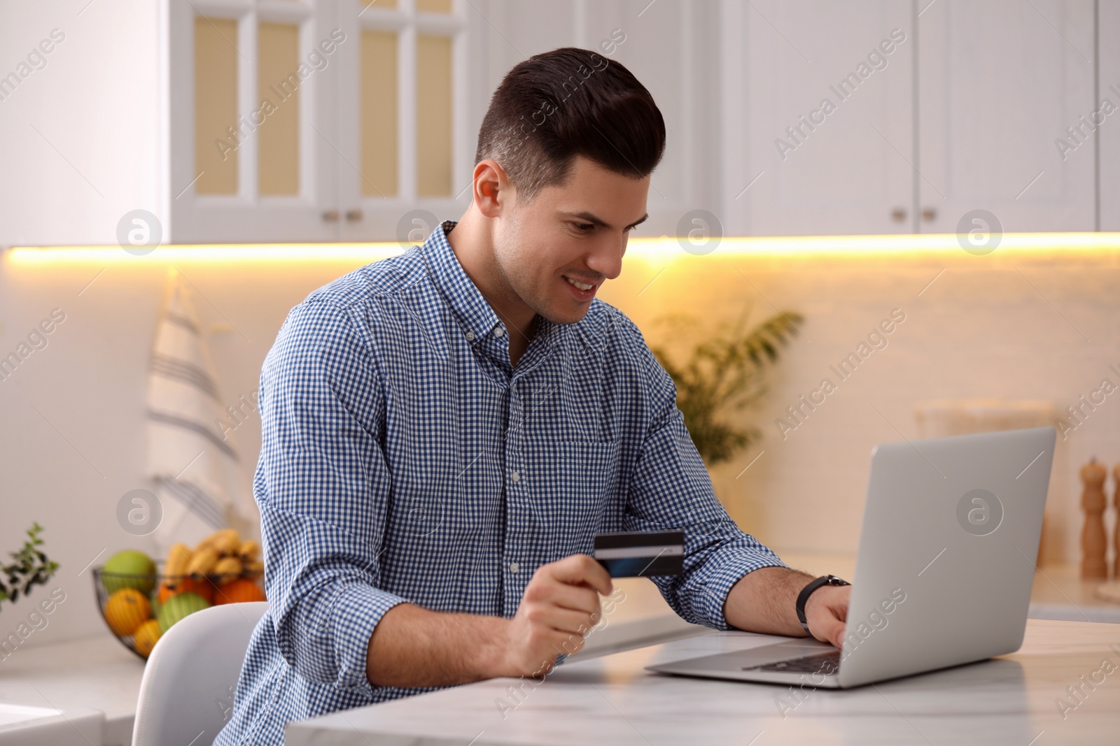 Photo of Man using laptop and credit card for online payment at table in kitchen