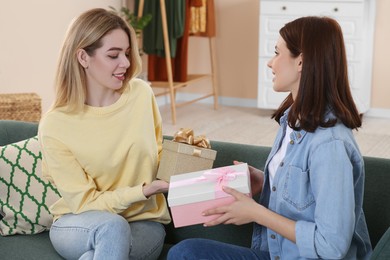 Photo of Smiling young women presenting gifts to each other on sofa at home