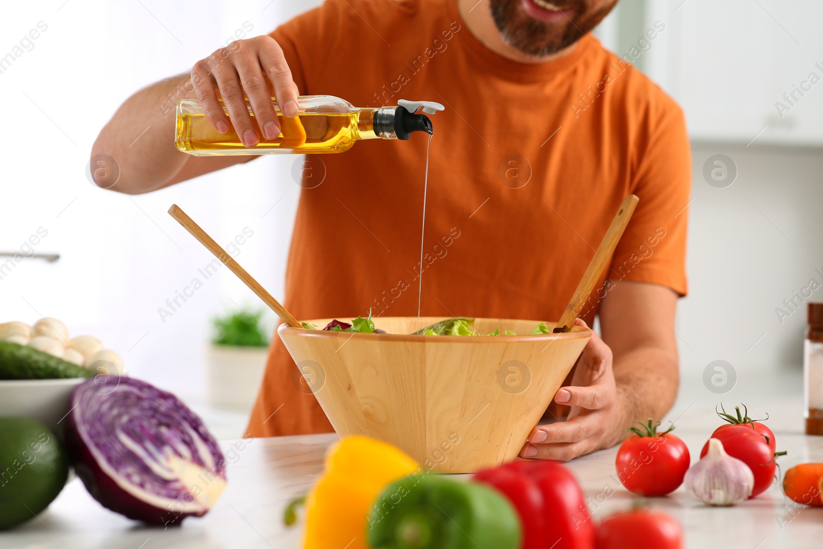 Photo of Man pouring oil into bowl of salad at table in kitchen, closeup. Online cooking course