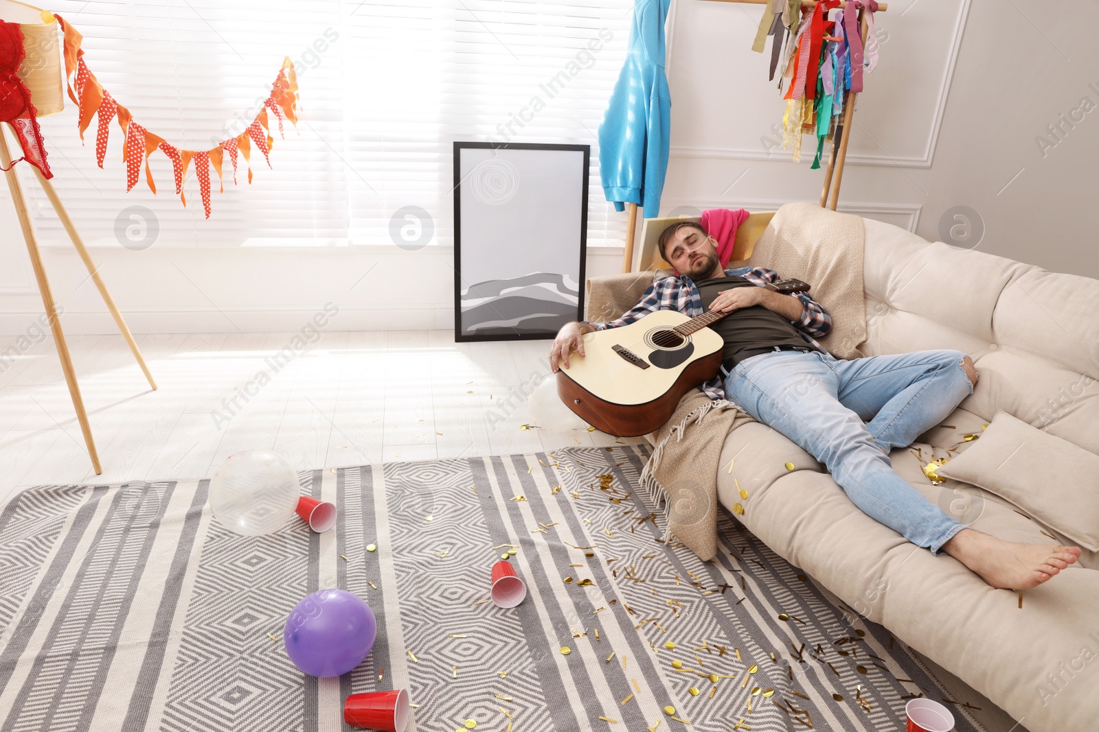 Photo of Young man with guitar sleeping on sofa in messy room after party