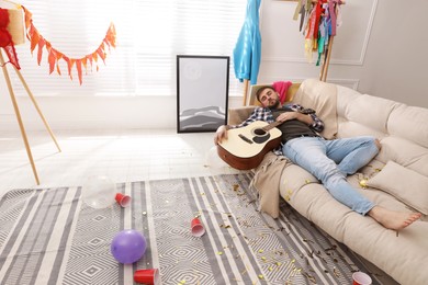 Photo of Young man with guitar sleeping on sofa in messy room after party