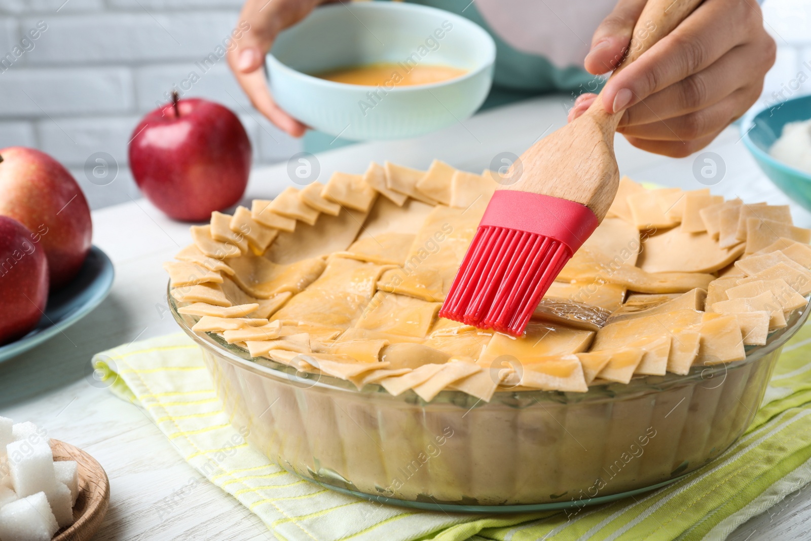 Photo of Woman spreading egg yolk onto raw apple pie at white wooden table, closeup