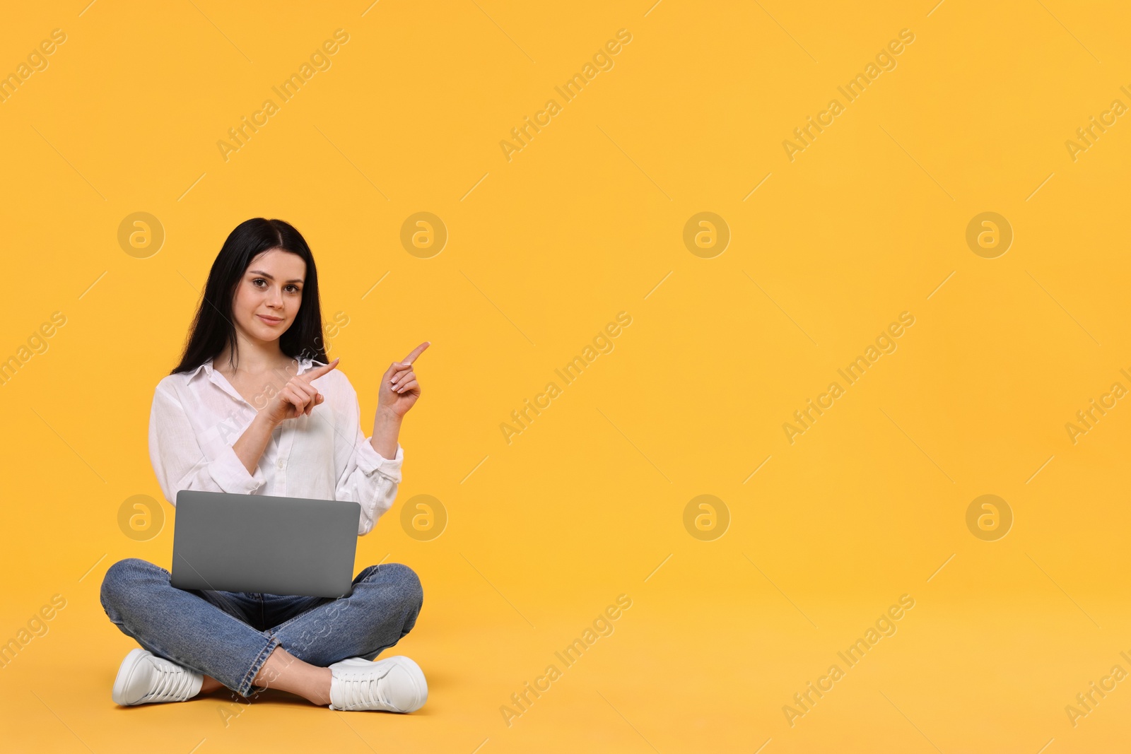 Photo of Student with laptop sitting and pointing at something on yellow background. Space for text