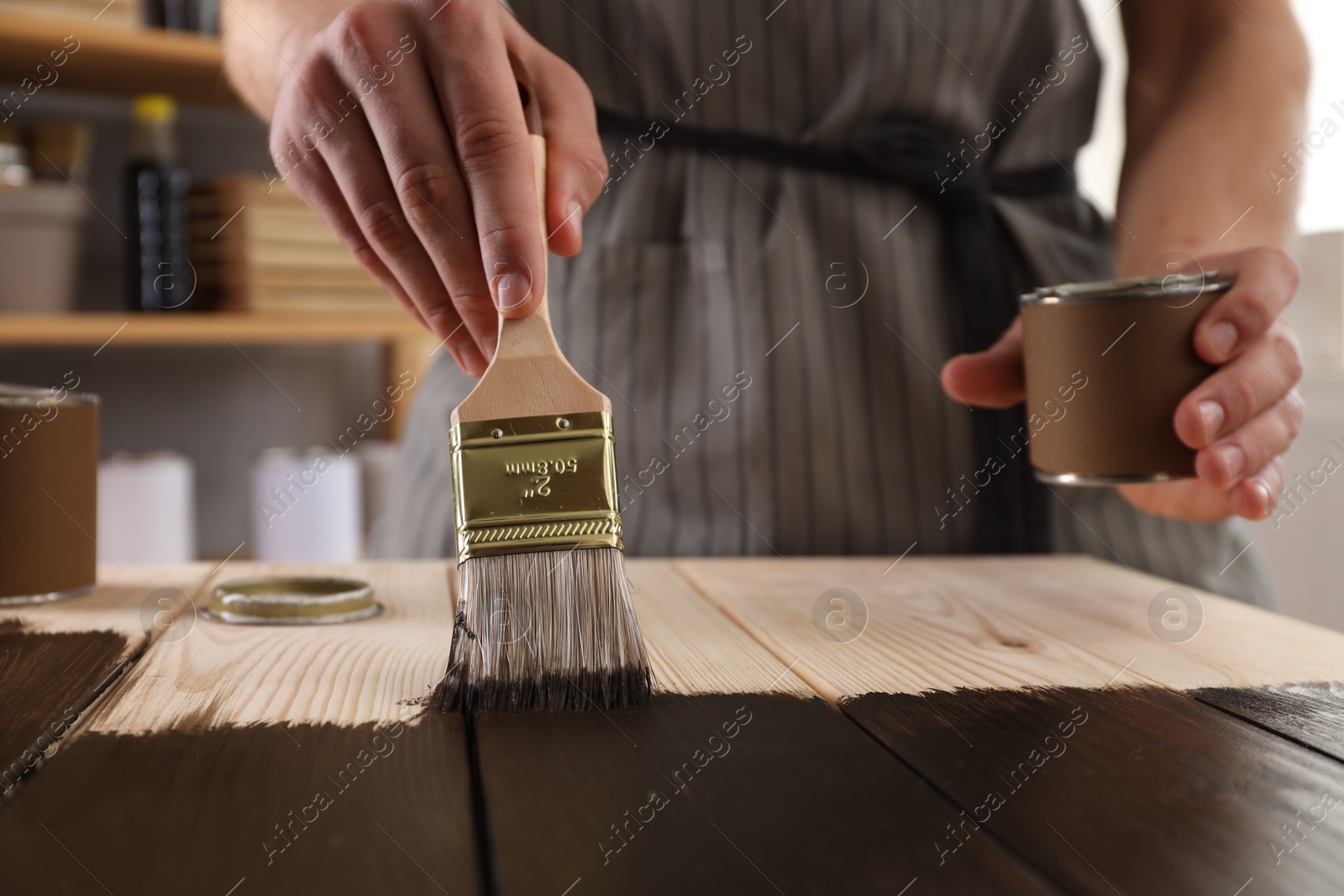 Photo of Man with brush and can applying wood stain onto wooden surface indoors, closeup