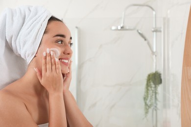 Photo of Beautiful young woman applying cleansing foam onto face in bathroom, space for text. Skin care cosmetic