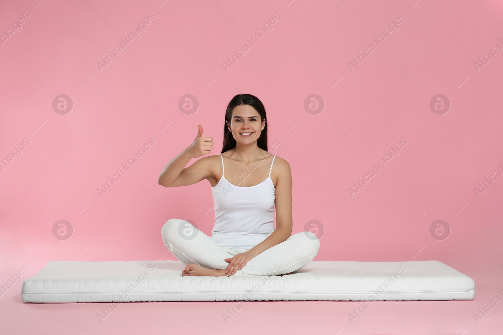 Photo of Young woman sitting on soft mattress and showing thumbs up against pink background