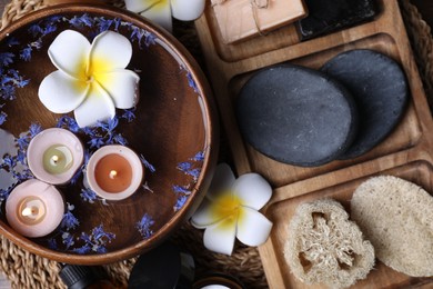 Photo of Different spa products, plumeria flowers and burning candles on wicker mat, top view