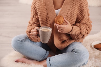Young woman with cup of coffee and cookie on rug at home, closeup. Winter atmosphere