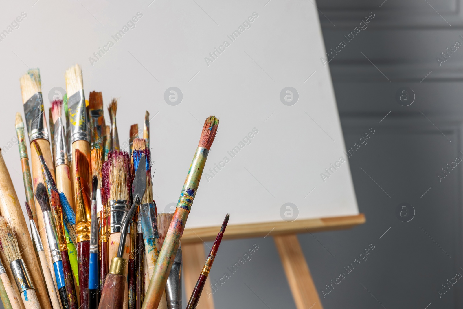 Photo of Easel with blank canvas and different art supplies near grey wall, closeup. Space for text