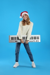Photo of Young woman in Santa hat with synthesizer on light blue background. Christmas music