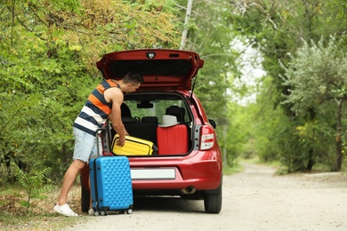 Happy man packing suitcases into car trunk near forest. Space for text