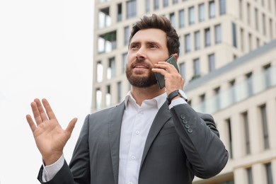 Handsome businessman talking on phone while walking near buildings outdoors