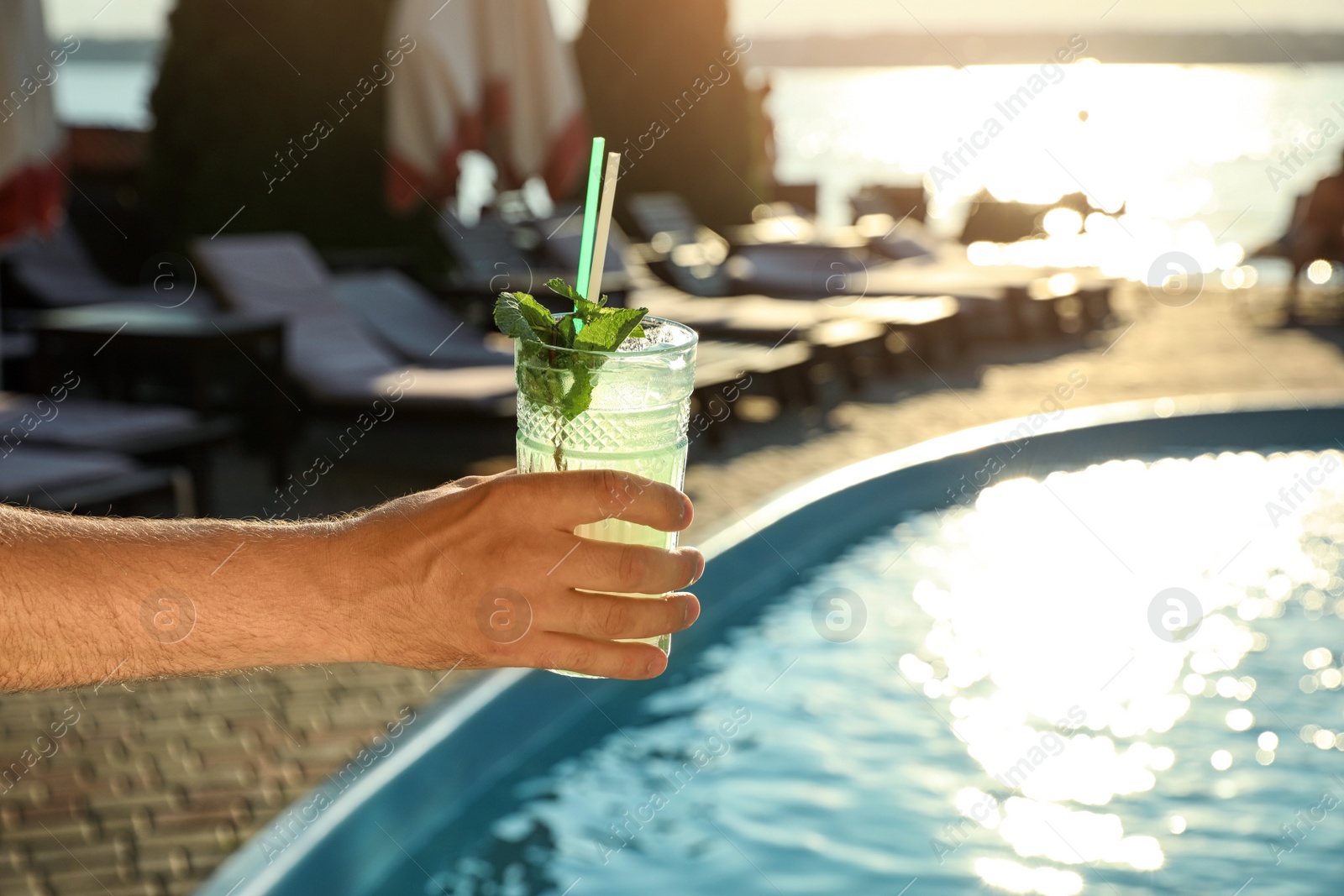 Photo of Man with glass of fresh summer cocktail near swimming pool outdoors at sunset, closeup. Space for text