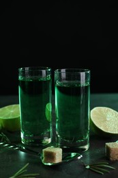 Photo of Absinthe in shot glasses, spoon, lime and brown sugar cubes on gray textured table, space for text. Alcoholic drink