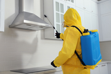 Photo of Pest control worker in protective suit spraying insecticide on furniture indoors. Space for text