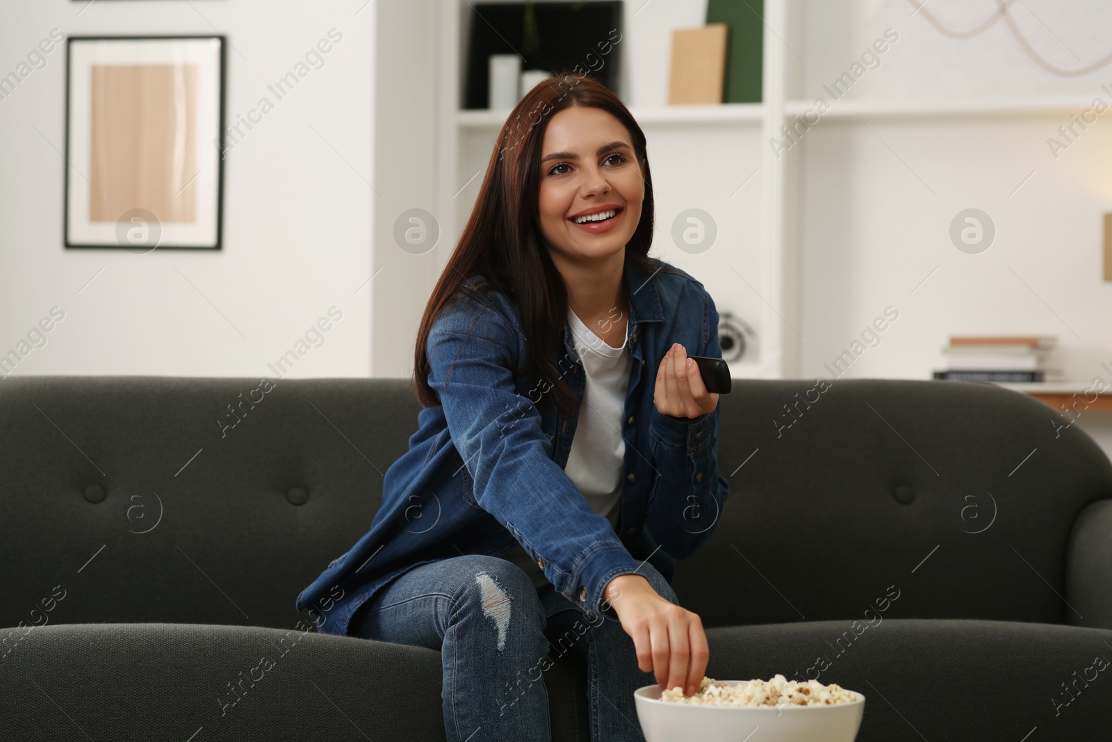 Photo of Happy woman changing TV channels with remote control on sofa at home