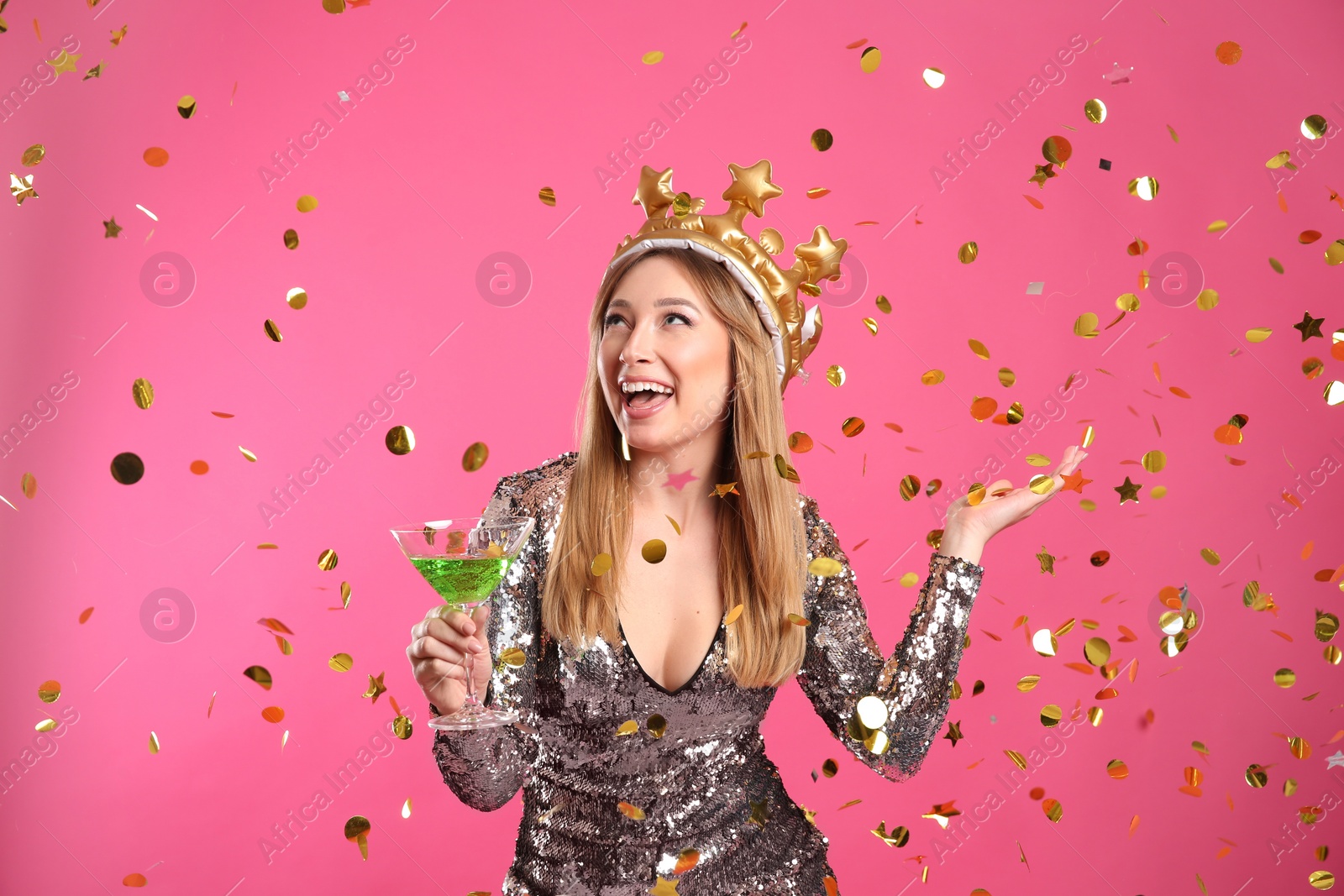 Photo of Happy young woman in party crown with cocktail and confetti on pink background