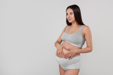 Photo of Beautiful pregnant woman in comfortable maternity underwear making heart with hands on her belly against grey background, space for text