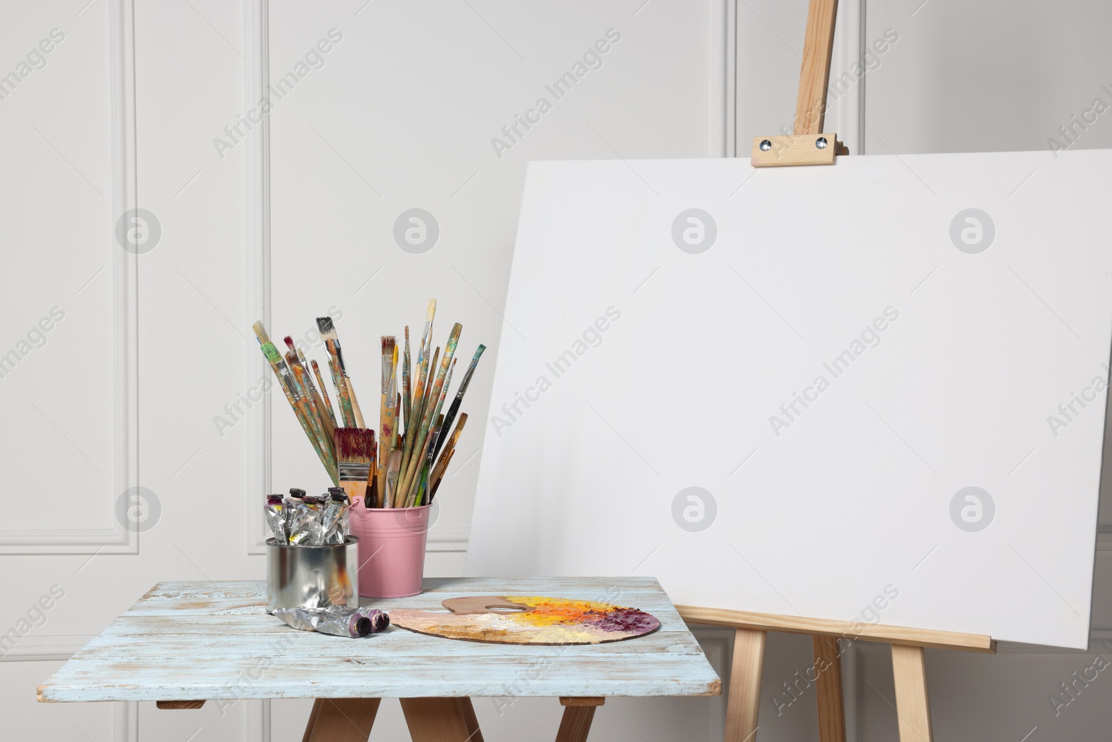 Photo of Easel with blank canvas and different art supplies on wooden table near white wall