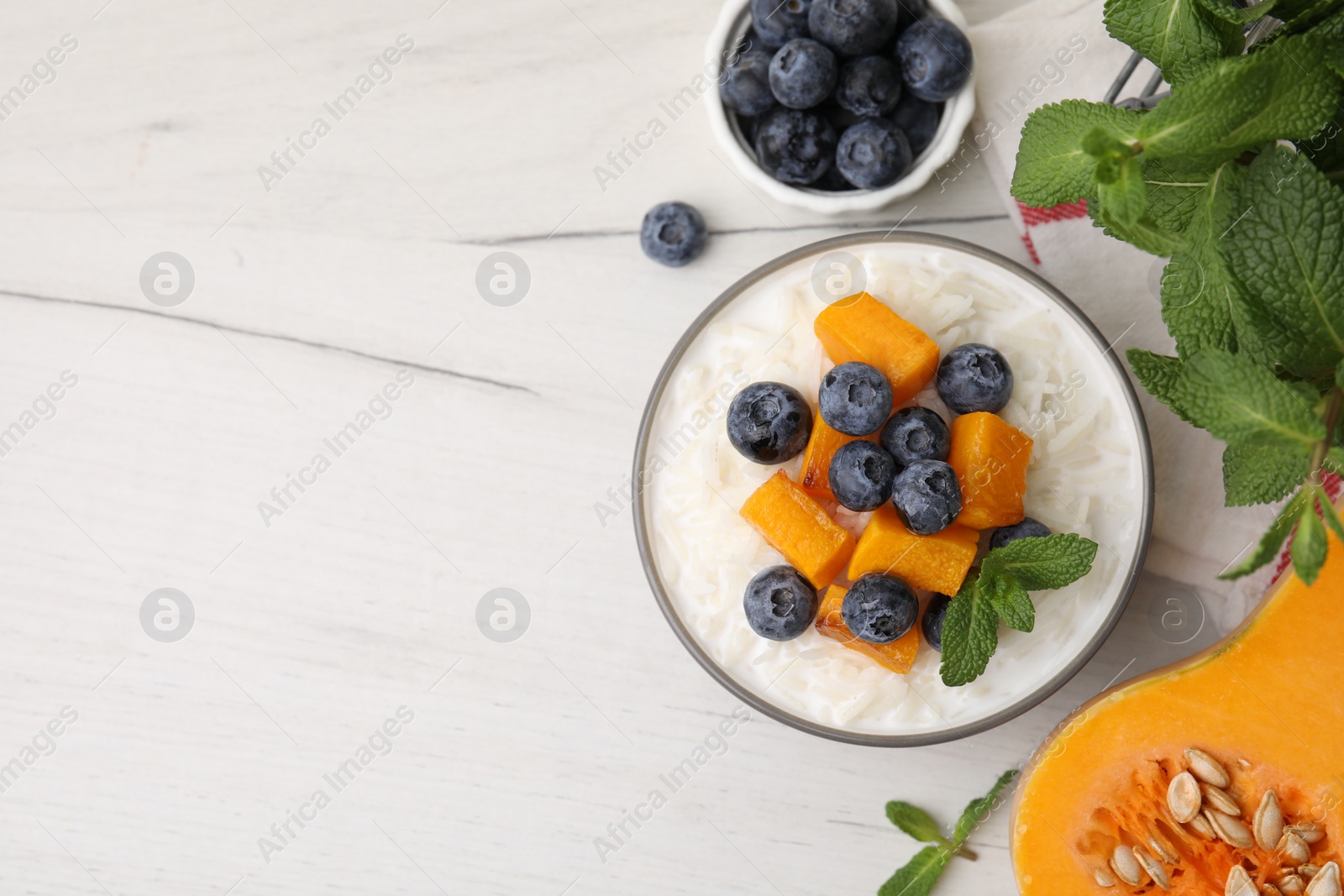 Photo of Bowl of delicious rice porridge with blueberries, pumpkin and mint on white table, flat lay. Space for text