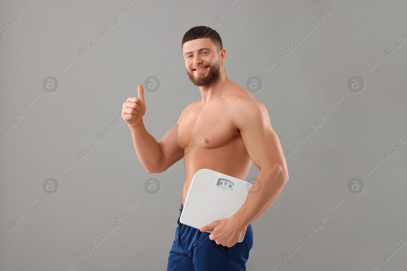 Photo of Happy athletic man holding scales and showing thumb up on grey background. Weight loss concept