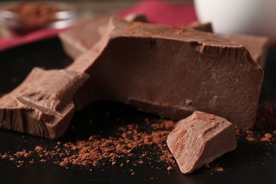 Photo of Pieces of tasty milk chocolate and cocoa powder on black plate, closeup