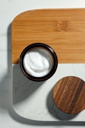 Photo of Board with open jar of cosmetic cream on white table, top view