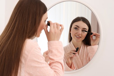 Beautiful young woman applying mascara near mirror indoors