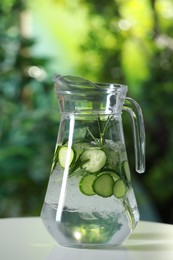 Photo of Refreshing cucumber water with rosemary in jug on white table against blurred green background