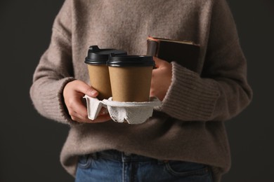 Woman holding takeaway cardboard cups and book on black background, closeup. Coffee to go