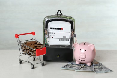Photo of Electricity meter, piggy bank, small shopping cart with coins and dollar banknotes on white wooden table